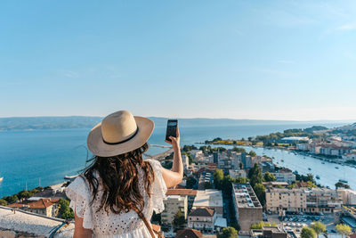 Rear view of woman using mobile phone, taking photos of beautiful seaside town of omiš in croatia