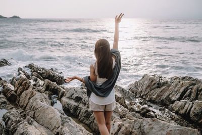 Rear view of woman waving hand while standing on rock by sea against sky