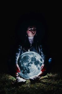 Young woman holding illuminated sphere while sitting on land at night