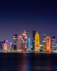 Sea by illuminated modern buildings against clear sky at night