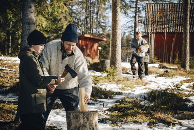 Father teaching son to cut log with axe in forest during winter