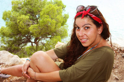 Caucasian girl smiles shyly sitting on a cliff with behind a blurred tree in summer 