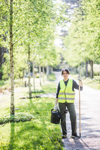 Full length portrait of confident young woman holding watering can and rake on footpath at garden