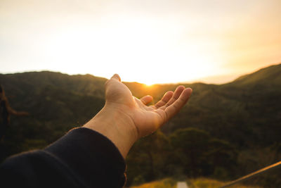 Cropped hand reaching mountains during sunset