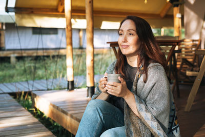 Young brunette woman in poncho drinking tea and relaxing in glamping in nature