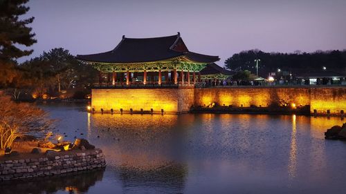 Illuminated building by lake against sky at dusk