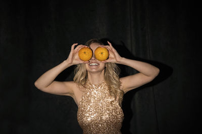 Playful woman covering eyes with oranges against black background