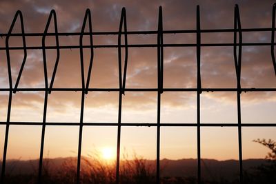 Silhouette metallic fence against cloudy sky during sunset