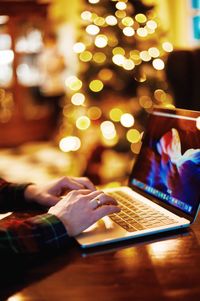 Cropped hands of man using laptop at table against illuminated christmas tree