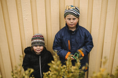 Portrait of brother and sister in warm clothing standing against wall in park