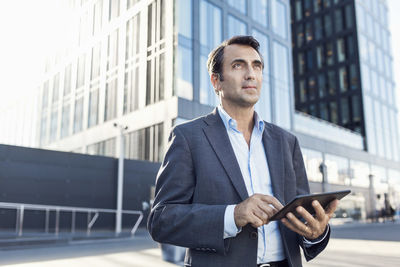 Confident businessman looking away while using digital tablet outside office building