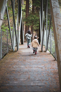 Sisters chase each other over a bridge on a forested path.