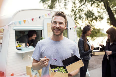 Smiling male customer holding disposable salad box against food truck with friends and owner in background