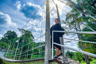 Portrait of young woman wearing graduation gown standing on footbridge at park