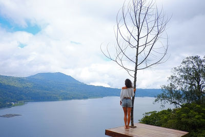 Woman standing by bare tree at lakeshore against sky