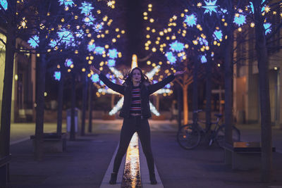 Portrait of woman standing on footpath against illuminated lights at night