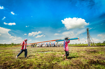 Side view of people carrying rocket while walking on grassy field against blue sky during sunny day