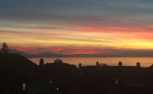 Silhouette buildings by sea against orange sky