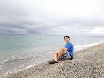 Young man sitting on shore at beach against sky