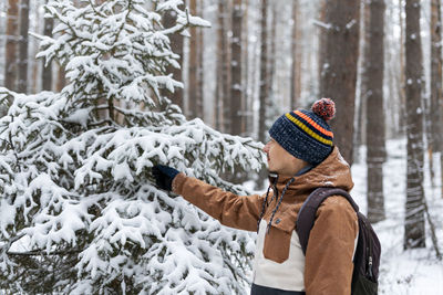 Young man in knitted hat walking in snowy forest in winter touching spruce branches enjoying  nature