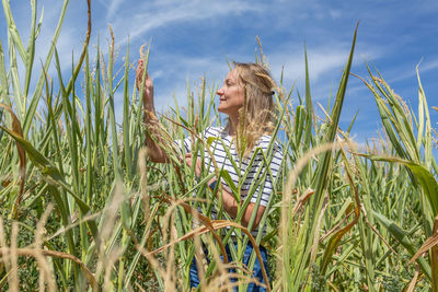 A woman in a corn field checks plants against a blue sky.