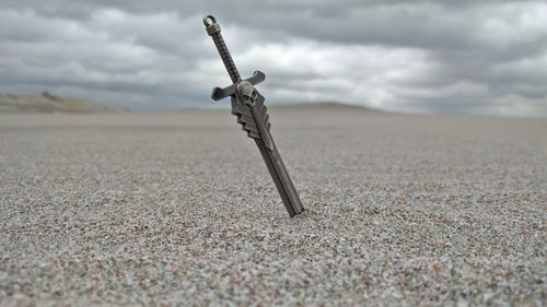 Sword in sand at beach against cloudy sky