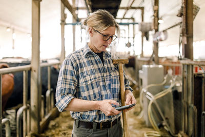 Farmer using smart phone standing with hammer at cattle farm