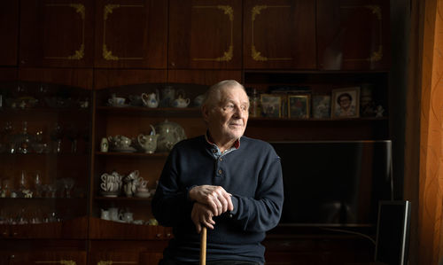 Thoughtful senior male in casual clothes with cane looking away with sad face while standing in living room at home