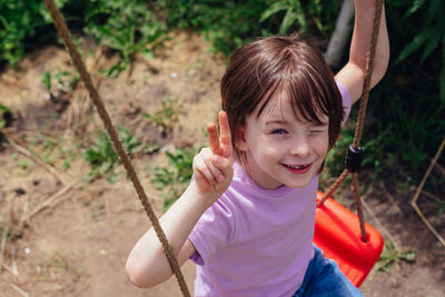 Portrait of a happy smiling child sitting and shows victory hand gesture on a swing on a sunny day. 