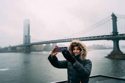 Smiling woman taking selfie while standing against manhattan bridge