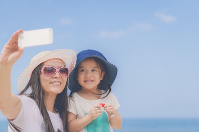 Happy mother and daughter wearing sunglasses enjoying at beach against sky