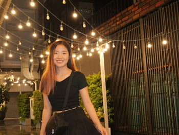 Portrait of young woman standing by illuminated string lights at night