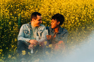 Loving young multiracial couple in casual clothes looking at each other and drinking champagne while sitting in lush blooming meadow during romantic date on sunny day