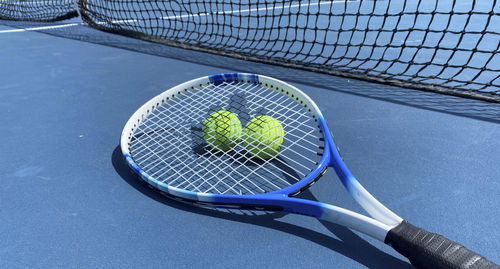 Horizontal view of two tennis balls with a racket on top on a blue tennis court.