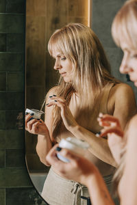 Blond woman holding moisturizer container in bathroom