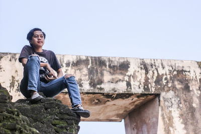 Low angle view of young man sitting on cliff against clear sky