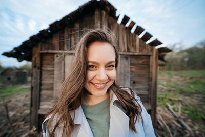 Beautiful girl with long hair in a grey trench coat next to an old wooden house outdoors in spring