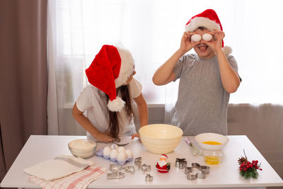 Brother and sister in red caps are having fun and preparing christmas cookies at a white table.