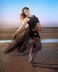 Pregnant woman standing at beach against sky during sunset