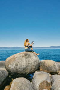 Man sitting on rock by sea against clear sky