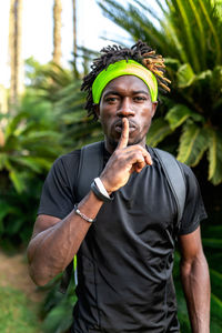 Young african american male athlete in sportswear and headband making shh gesture while standing against green tropical plants on street