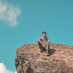 Young man sitting on rock against blue sky