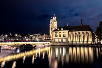 Bridge over river by illuminated buildings against sky at night