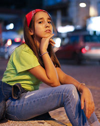 Young woman looking away while sitting on street in city at night