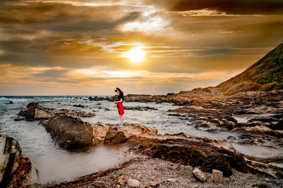 Side view of girl standing on rock at beach against sky during sunset