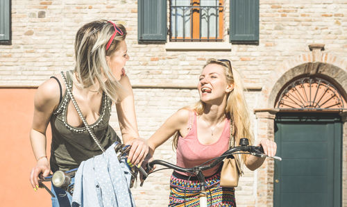 Cheerful female friends riding bicycles against building