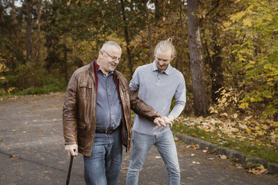 Young male caretaker supporting retired senior man walking with cane on road