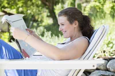 Side view of smiling woman reading newspaper while relaxing on lounge chair at yard