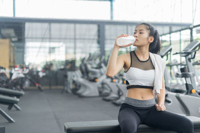 Young woman drinking water from bottle while sitting at gym