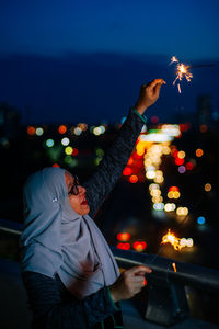 Side view of smiling mid adult woman holding burning sparkler at night